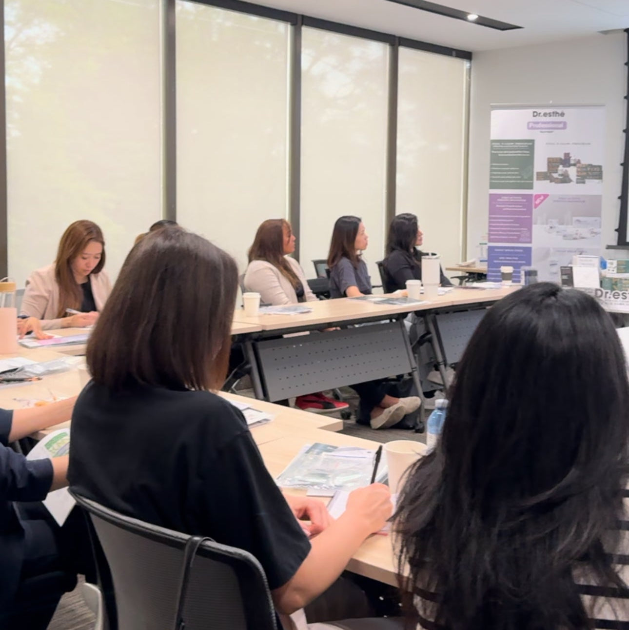 Group of people sitting at tables in a conference room, learning professional Korean skincare with large windows