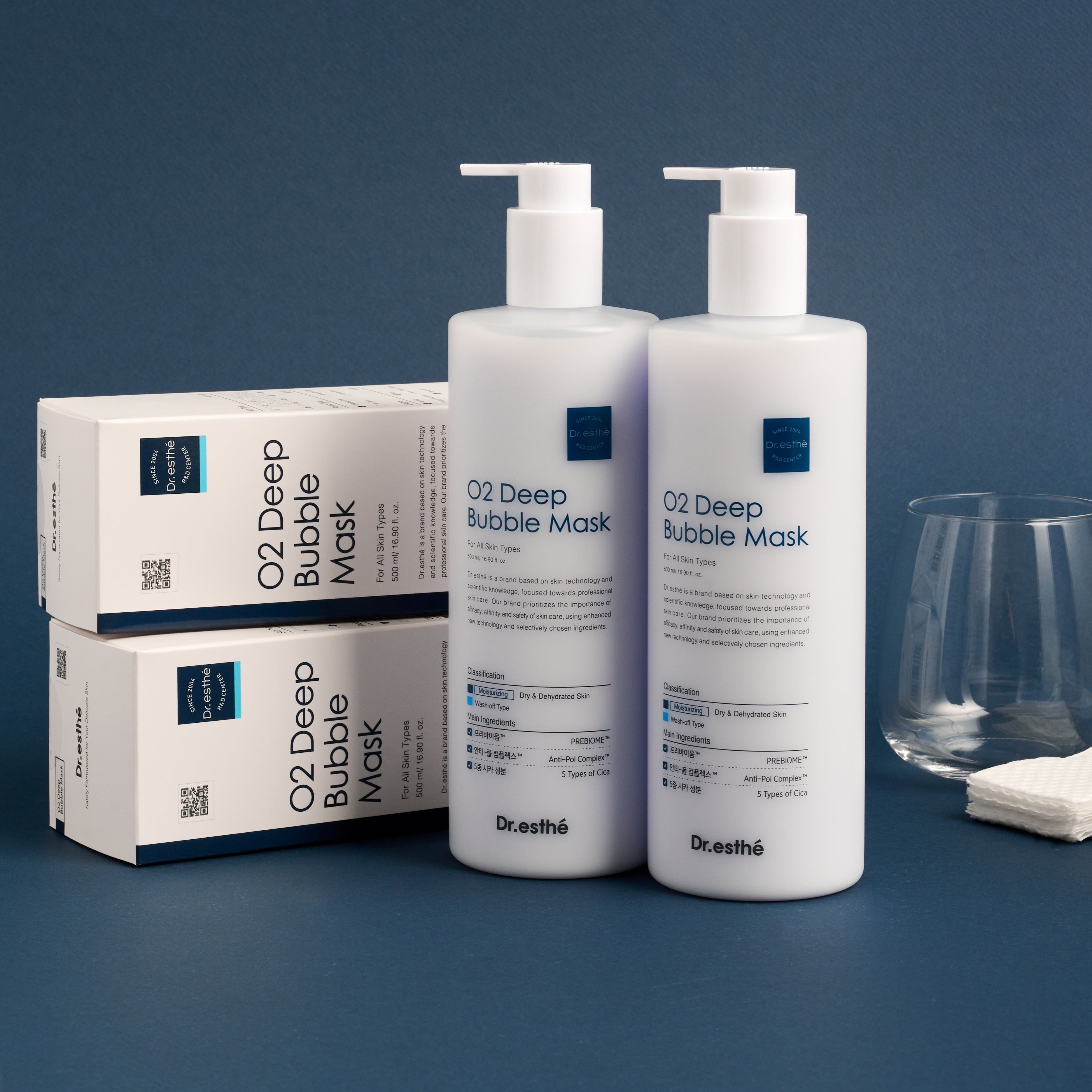Two bottles of Dr. Esthe O2 Deep Bubble Mask on a blue background with boxes and a glass.